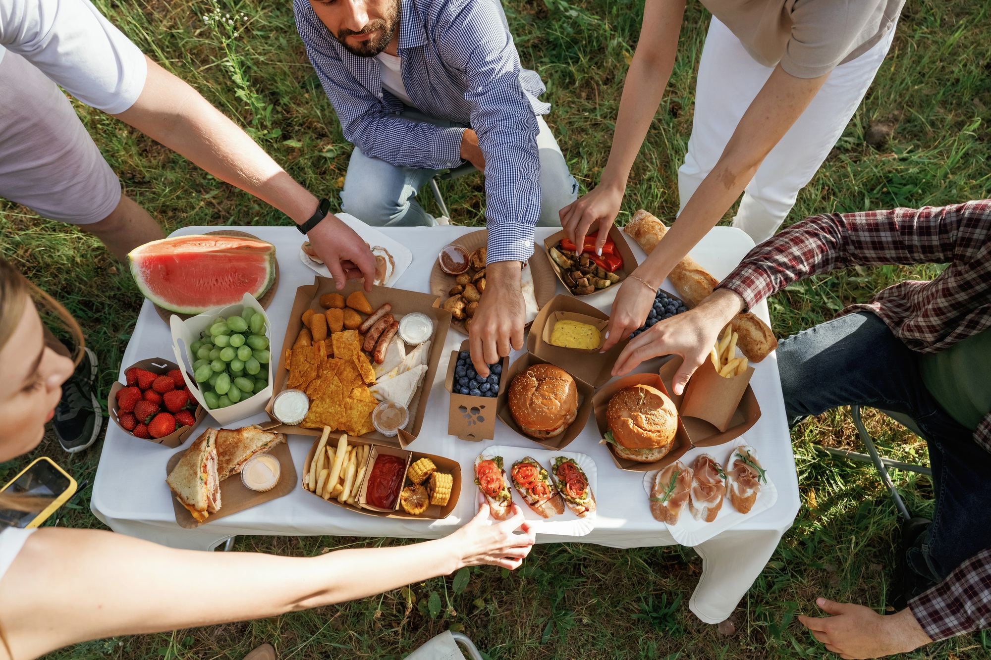 Meal is on the table. Group of friends are having picnic on the field with food in eco boxes