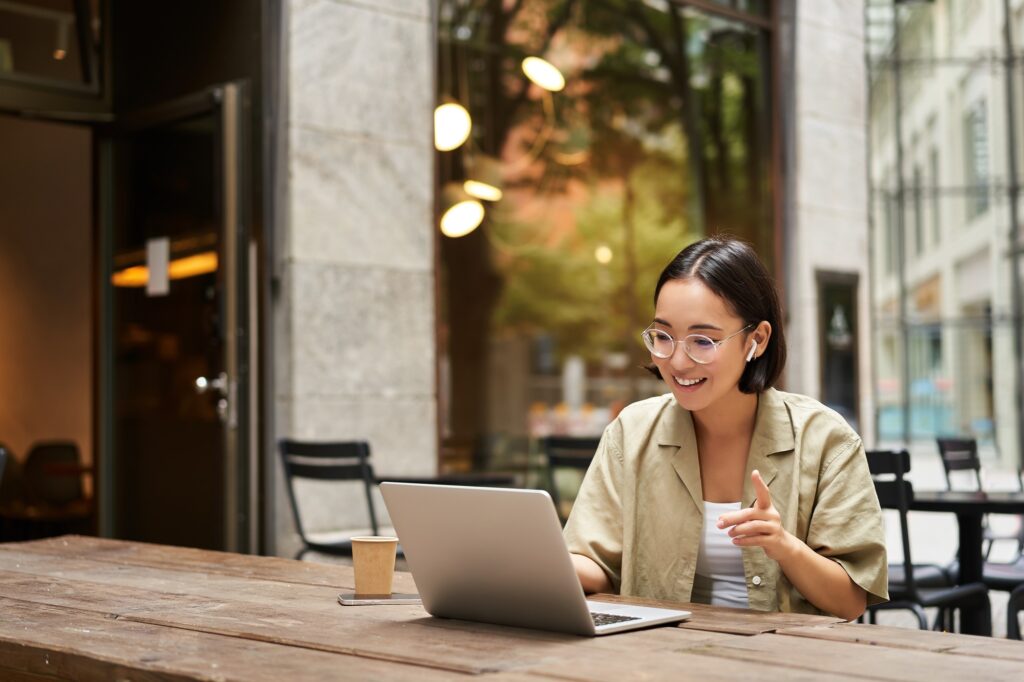 Young woman sitting on online meeting in outdoor cafe, talking to laptop camera, explaining